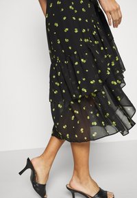 Femme marchant dans une jupe midi noire transparente avec un petit imprimé floral jaune et des sandales noires à talons ouvertes à l'arrière.