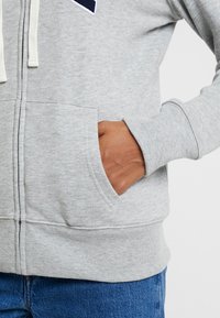 Mano parcialmente dentro del bolsillo frontal de una sudadera con cremallera gris claro, llevada con jeans azules, contra un fondo sencillo.