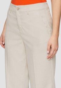 Beige wijde broek met een gladde textuur, enkele knoopsluiting en zijzakken. Zichtbare stikseldetails bij de naden.