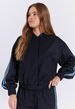 Femme aux longs cheveux bruns portant un survêtement noir avec des bandes bleu clair sur les manches et le pantalon, debout, une main sur la hanche.