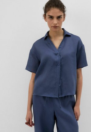 Jonge vrouw draagt een loszittend navy blauw overhemd met korte mouwen en bijpassende broek, staand tegen een effen lichte achtergrond.