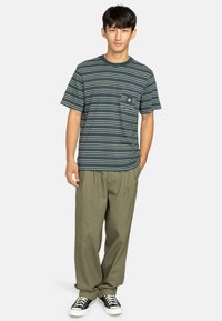 Striped t-shirt in navy blue with green and white lines, featuring a front pocket. Paired with loose-fitting olive green pants and black sneakers.