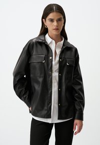 Giacca in pelle sintetica nera con colletto, due tasche sul petto e bottoni a pressione argentati, indossata sopra una camicia bianca.