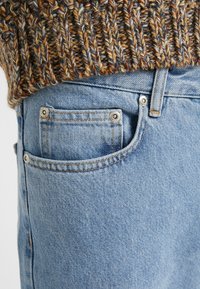 Vaqueros de denim azul claro con un bolsillo frontal que presenta remaches plateados. La parte superior es un suéter de punto texturizado en tonos tierra.