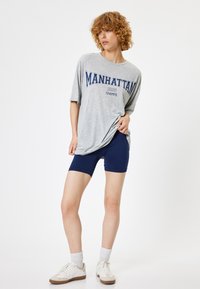Szara, oversize'owa koszulka z nadrukiem "MANHATTAN 1995 CHAMPS", połączona z ciemnoniebieskimi szortami rowerowymi i białymi sneakersami. Wykonana z bawełny, o swobodnym kroju.