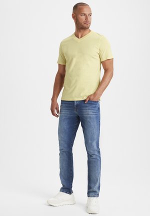 Helles gelbes T-Shirt mit kurzen Ärmeln und V-Ausschnitt, kombiniert mit blauen Slim-Fit-Jeans und weißen Sneakers. Einfaches Design, lässiger Look.