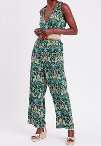 Combinaison à motifs avec un décolleté en V profond, jambes larges, en vert, bleu et noir, avec une ceinture tissée. Portée avec des sandales compensées beige.