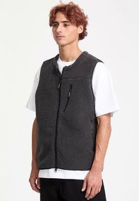 Gilet en polaire gris à texture douce, avec une fermeture éclair sur toute la longueur et deux poches frontales, porté par-dessus un t-shirt blanc. Design décontracté et fonctionnel.