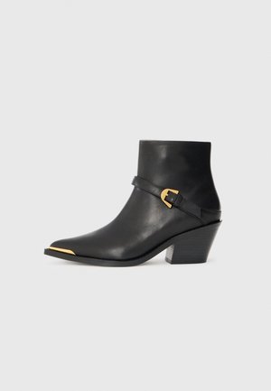 Bottine en cuir noir avec un bout pointu, accent doré à la pointe et une sangle avec une boucle. Présente un talon épais et une texture lisse.