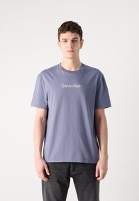 Gai violetas kokvilnas T-krekls ar īsām piedurknēm, kuram ir apaļa kakla izgriezums un baltais "Calvin Klein" logotips priekšā. Saskaņots ar tumšām džinsēm.