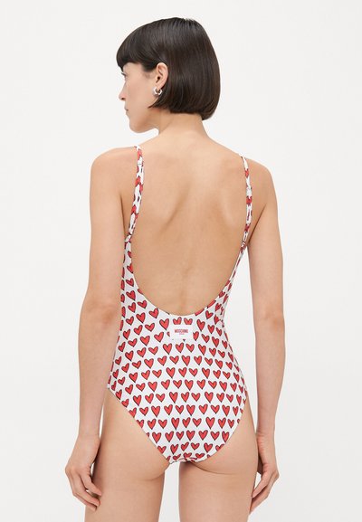 One-piece swimsuit with red and light blue heart pattern on white fabric. Low back design, thin straps, and visible logo label at the back.