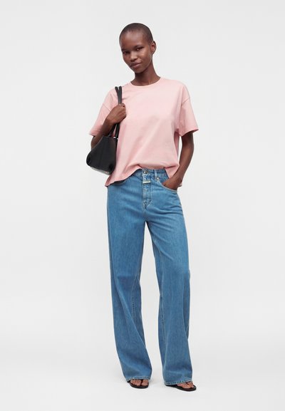 Personne debout portant un t-shirt rose pâle, un jean bleu à jambes larges, des sandales noires et portant un sac à bandoulière noir contre un fond uni.