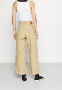 Béžové cargo kalhoty s širokými nohavicemi, dvojitými zadními kapsami, nášivkou na pase a ohrnutými lemy, v kombinaci s černými vysokými teniskami.