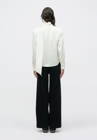 Kremowa jedwabna bluzka z wiązaniem z tyłu, długimi rękawami i zaokrąglonym dołem, w połączeniu z szerokimi czarnymi spodniami, widziana od tyłu.