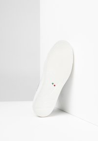 Suela de zapato de goma blanca con agarre texturizado, que presenta un pequeño acento de la bandera italiana y detalles del logotipo en relieve, colocada en ángulo sobre un fondo liso.