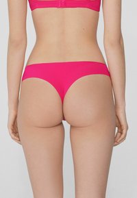 Bañador de bikini rosa con una textura suave y elástica, que presenta un diseño de cintura baja y cobertura mínima, acentuando la forma del cuerpo.