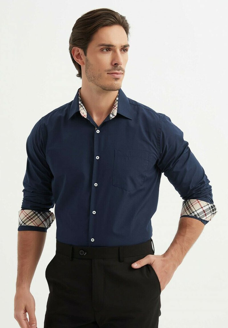 Chemise bleu marine à boutons avec une poche unique sur la poitrine et des poignets en tartan. Le tissu semble lisse ; le col présente un motif tartan en contraste.