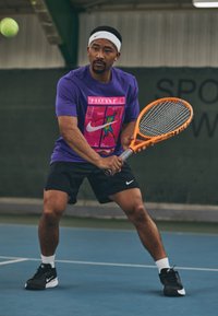 Άνδρας με μωβ πουκάμισο και μαύρη βερμούδα ετοιμάζεται να χτυπήσει τένις με πορτοκαλί ρακέτα σε κλειστό γήπεδο.