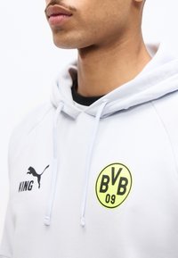 Detail muže v bílém kapuci s logem Borussia Dortmund (BVB 09) a logem Puma, na hrudi s nápisem "KING".