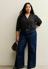 Woman wearing black button-up blouse and wide-leg dark blue jeans, holding black handbag, standing against plain light wall.