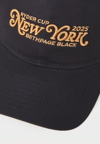 Crna prilagođena kapa s zlatnim vezenim tekstom: "RYDER CUP NEW YORK 2025 BETHPAGE BLACK." Ima zakrivljeni obod i teksturiranu tkaninu.