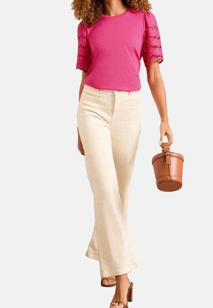 Frau trägt ein rosa Hemd mit Spitzenärmeln, cremefarbene Hosen mit weitem Bein, braune Sandalen mit Absatz und hält eine braune Leder-Bucket-Bag.