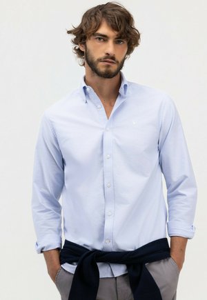 Hombre con cabello castaño y barba que lleva una camisa azul claro con botones y las mangas remangadas, y un suéter oscuro atado alrededor de su cintura.