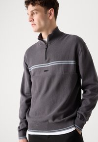 Sivá zip-up kapuca z teksturirane tkanine, ki ima horizontalno modro črto in logo. Dolgi rokavi z rebrastimi manšetami in robom.
