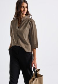 Chemise boutonnée vert olive avec col et manches trois-quarts, associée à un pantalon noir ; tenant un sac à main beige. Texture lisse.