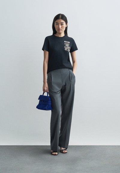 Γυναίκα φορώντας μαύρο μπλουζάκι Moschino με γραφική απεικόνιση αρκούδας, γκρι κομψά παντελόνια, μαύρες σαγιονάρες, κρατώντας μια μικρή μπλε τσάντα, στέκεται μπροστά από λευκό φόντο.