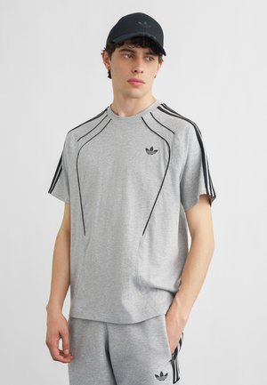 Ung mand iført gråt Adidas træningssæt og sort kasket, stående med den ene hånd i lommen og kigger til siden.