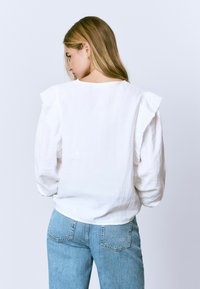 Blusa blanca texturizada con detalles de hombros con volantes, combinada con jeans de color azul claro. Escote sencillo y corte holgado, mostrando un diseño casual.