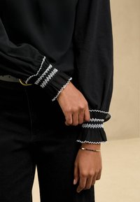 Schwarze Bluse mit langen Ärmeln und weißen bestickten Manschetten, die mit Falten und gewellten Kanten verziert ist, kombiniert mit schwarzen Jeans und einem goldenen Armband.