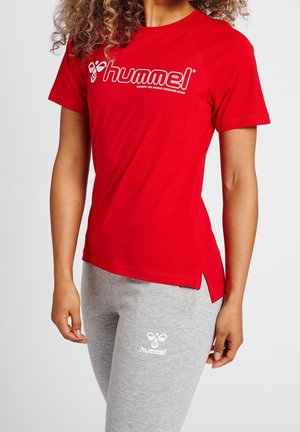 Czerwona koszulka krótki rękaw z białym logo "hummel". Posiada boczne rozcięcia u dołu. Połączona z szarymi spodniami dresowymi, również znakowanymi logo.