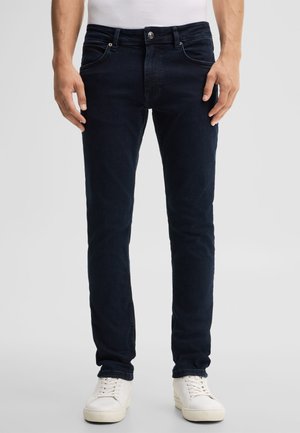 Man wearing dark blue slim-fit jeans and white sneakers standing against a plain light background.