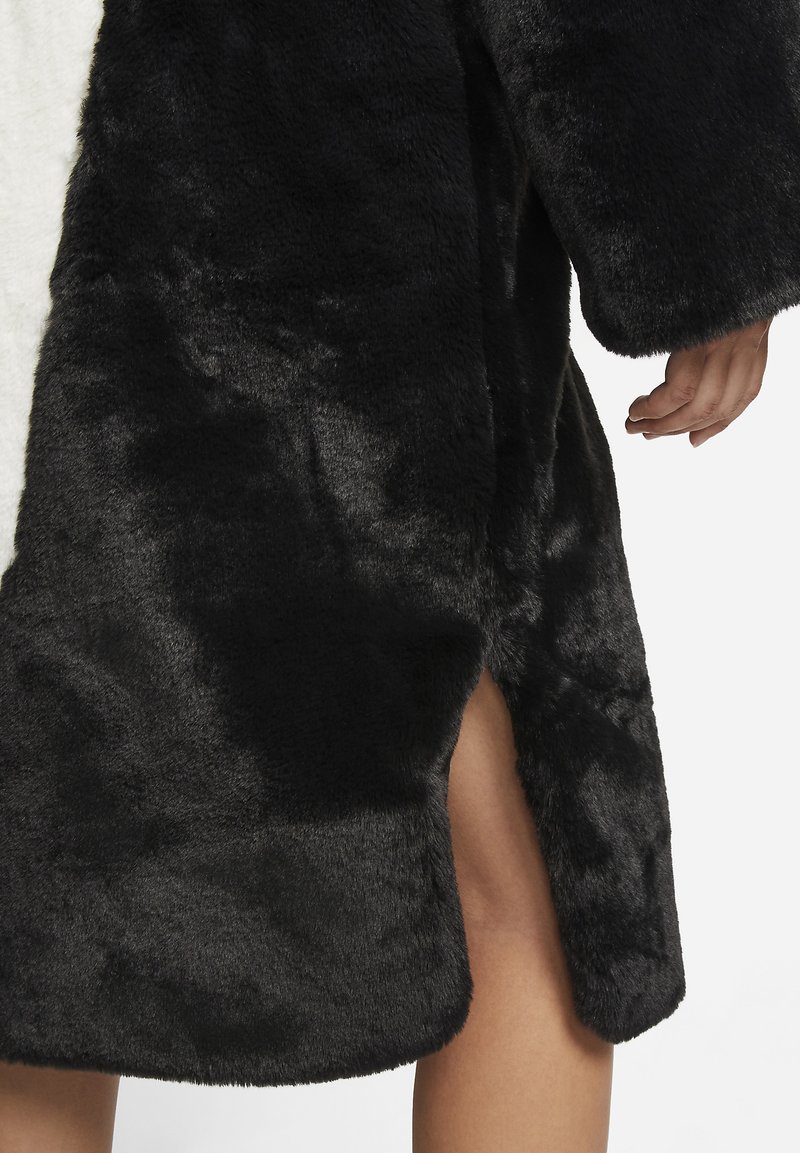 Abrigo de piel sintética negra con un acabado suave y texturizado, que presenta una abertura lateral y mangas anchas para un ajuste oversize.