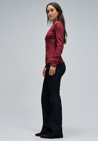 Top de manga larga de canalé en color burdeos con detalles de botones, combinado con jeans acampanados oscuros y zapatos marrones, sobre un fondo gris claro.