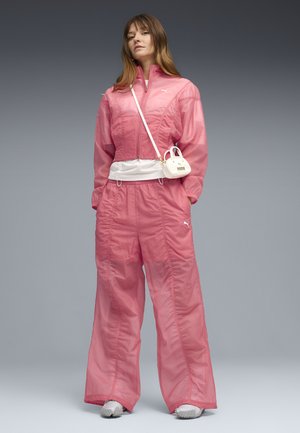 Femme portant un survêtement rose Puma, des baskets blanches et un petit sac blanc porté en bandoulière sur un fond dégradé gris.