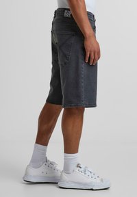 Dunkelgraue Denim-Shorts mit geradem Schnitt, ausgestattet mit Gesäßtaschen und gelben Nahtakzenten, kombiniert mit weißen Sneakern und Socken.