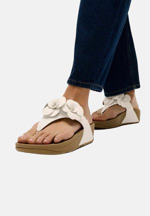 Pieds portant des sandales compensées blanches avec de grandes décorations florales, associées à un jean denim bleu foncé.
