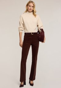Beżowy sweter z dzianiny z wysokim kołnierzem, brązowe szerokie spodnie sztruksowe, pasek ze złotą klamrą i bordowa torebka. Dopełnieniem stylizacji są buty na wysokim obcasie.