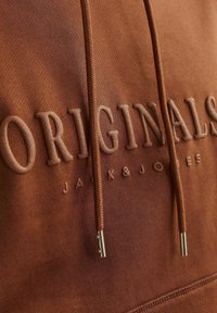 Brun hættetrøje med "ORIGINALS" og "JACK & JONES" prægede i en matchende farve. Har snøre med silder metalspidser og blød stoftekstur.