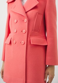 Abrigo de lana en color rosa coral con diseño de doble botonadura, que presenta seis botones, dos bolsillos de solapa y un gran cuello con muescas.