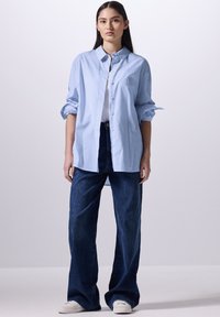 Camicia azzurra chiara con maniche arrotolate, abbinata a jeans larghi blu scuro e sneakers bianche. Texture di cotone liscia, design classico.