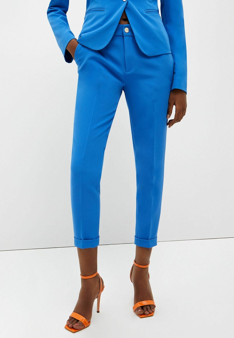 Femme portant un pantalon bleu vif sur mesure longueur cheville et un blazer assorti, associé à des sandales orange à talons hauts.