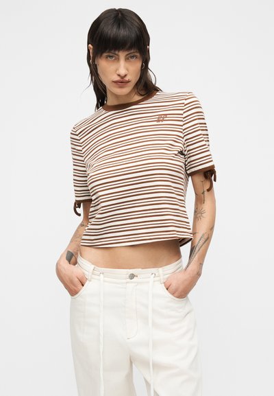 Γυναίκα με σκούρα μαλλιά που φοράει ριγέ καφέ και λευκό κοντομάνικο τοπ και λευκό παντελόνι, στέκεται με τα χέρια στις τσέπες μπροστά σε λευκό φόντο.