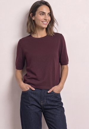 Femme aux cheveux mi-longs portant un haut bordeaux à manches courtes et un jean bleu foncé, debout les mains dans les poches, souriante.