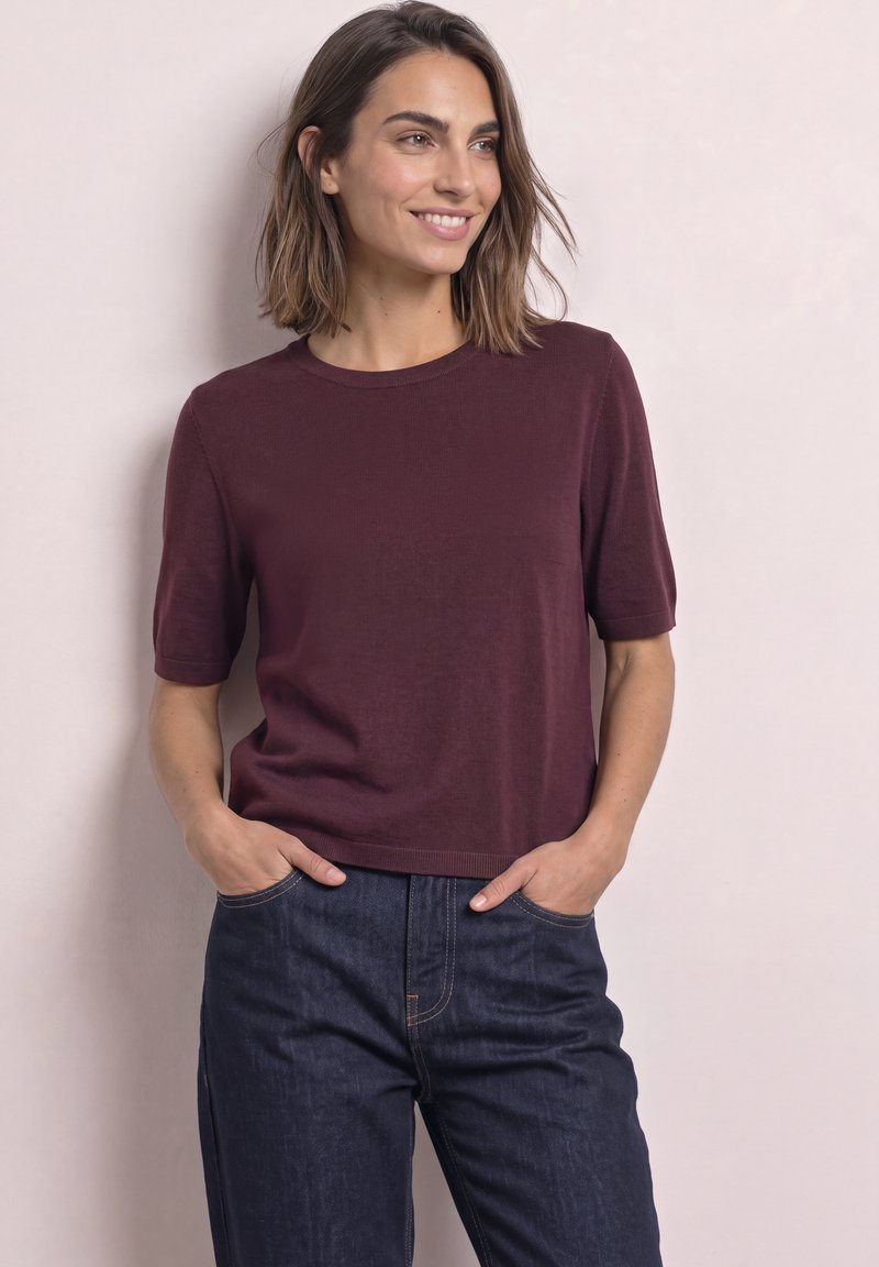 Femme aux cheveux mi-longs portant un haut bordeaux à manches courtes et un jean bleu foncé, debout les mains dans les poches, souriante.