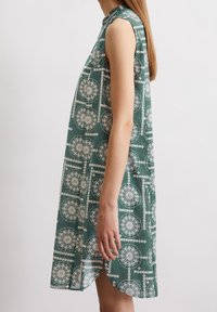 Grön ärmlös klänning med vitt cirkelmönster och botaniska detaljer. Tyget har en mjuk textur och en avslappnad passform.