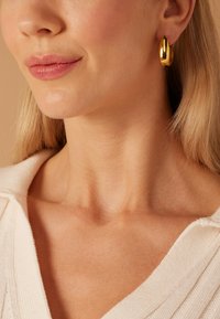 Gouden oorringen met een gladde, reflecterende oppervlakte, gevormd als verlengde druppels. Minimale textuur, wat perfect past bij een top in neutrale tinten.